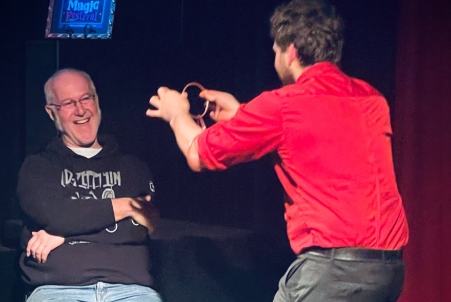 A magician hold a ring in front of a man who is seated and laughing. 