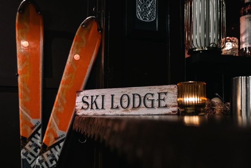 Two vintage orange skis resting against a bar counter with a white wooden panel sign on the bar that reads SKI LODGE.