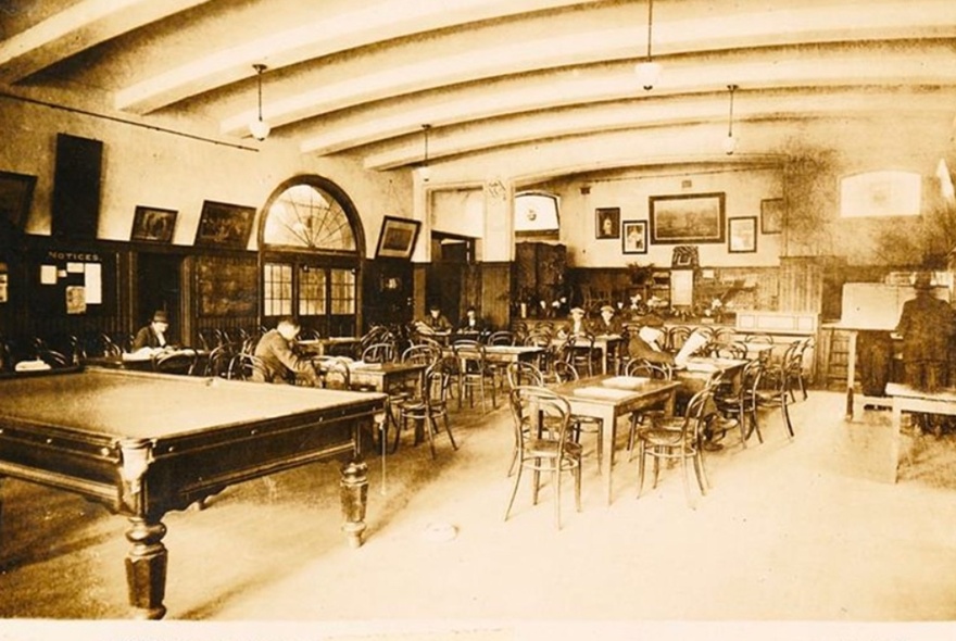 Vintage sepia image of a large hall at the Mission to Seafarers building with a pool table, table and chairs, paintings on the walls, and a curved ceiling.