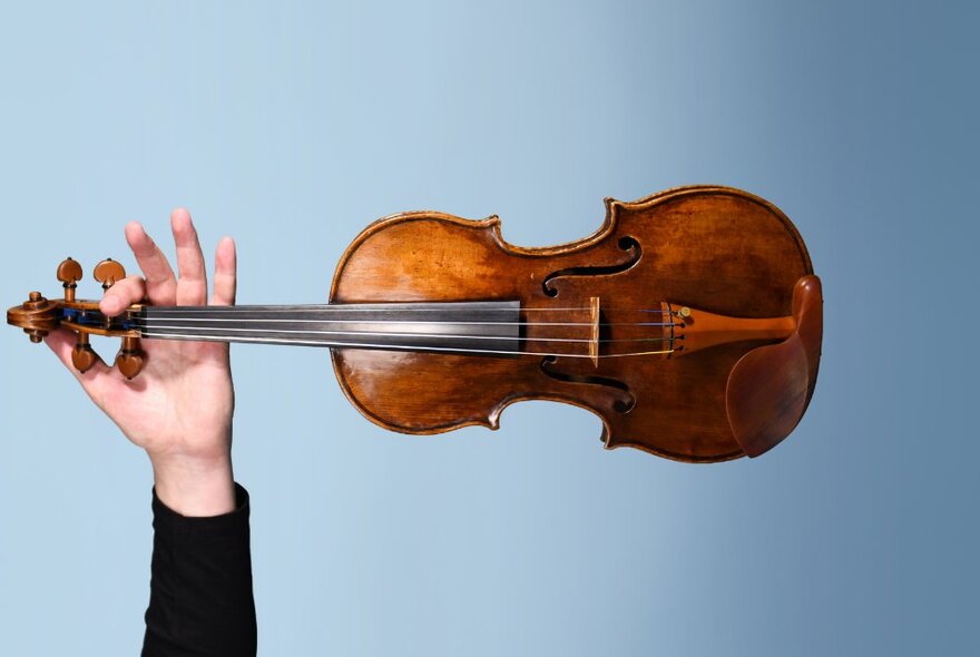 A violin being held in a horizontal position by a raised hand, against a pale blue background.