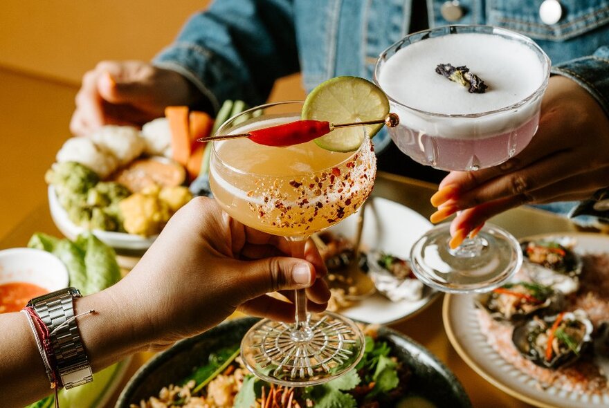 People clinking glasses of different cocktails over a table set with many plates of food. 