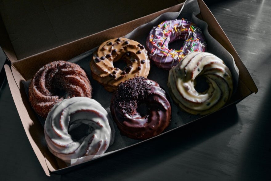 A take-away box of six crullers, all with different flavoured icing.