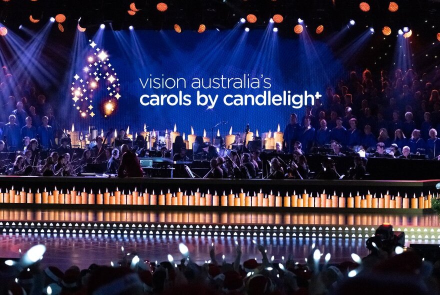 Crowded stage with massed row of candles, choir, orchestra and signage under lights.