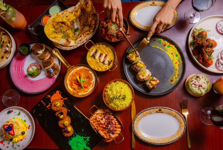 Hands holding cutlery slicing into a kebab on a table laden with small dishes of brightly coloured Indian food.