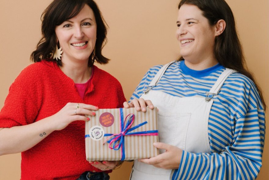 Two smiling people standing side by side, holding a wrapped gift between them.