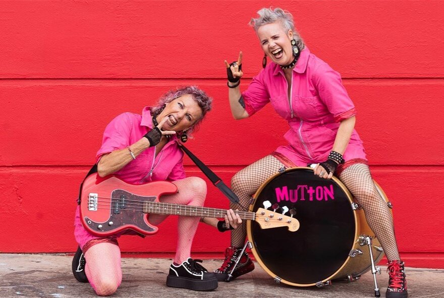 Two older women with guitar and drum, wearing pink outfits and laughing.