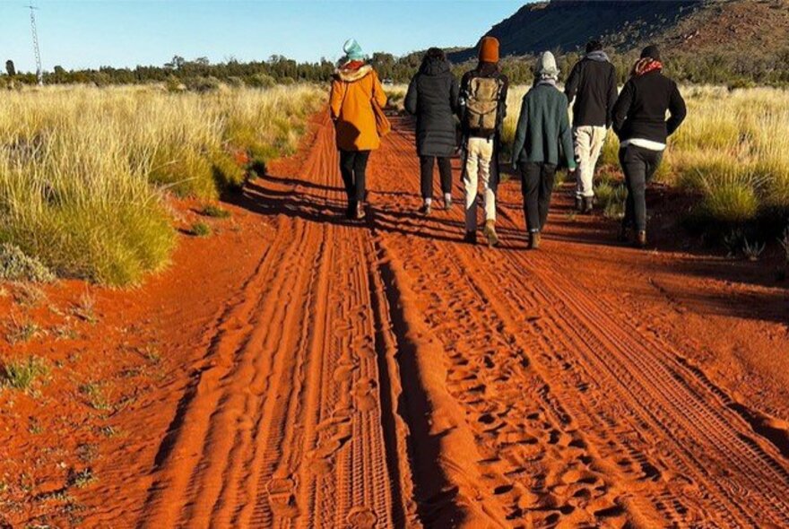 People walking along a red sand road in the outback and fields of dry grass.
