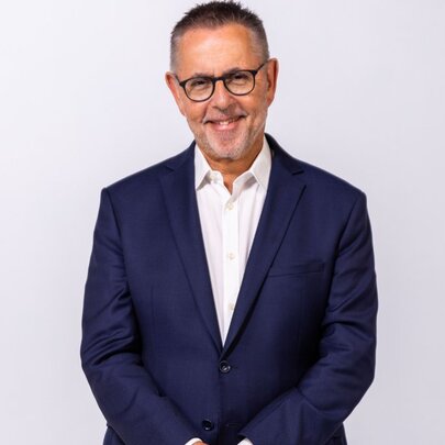 Dr Norman Swan, smiling warmly as he poses against a white wall wearing a blue jacket. 