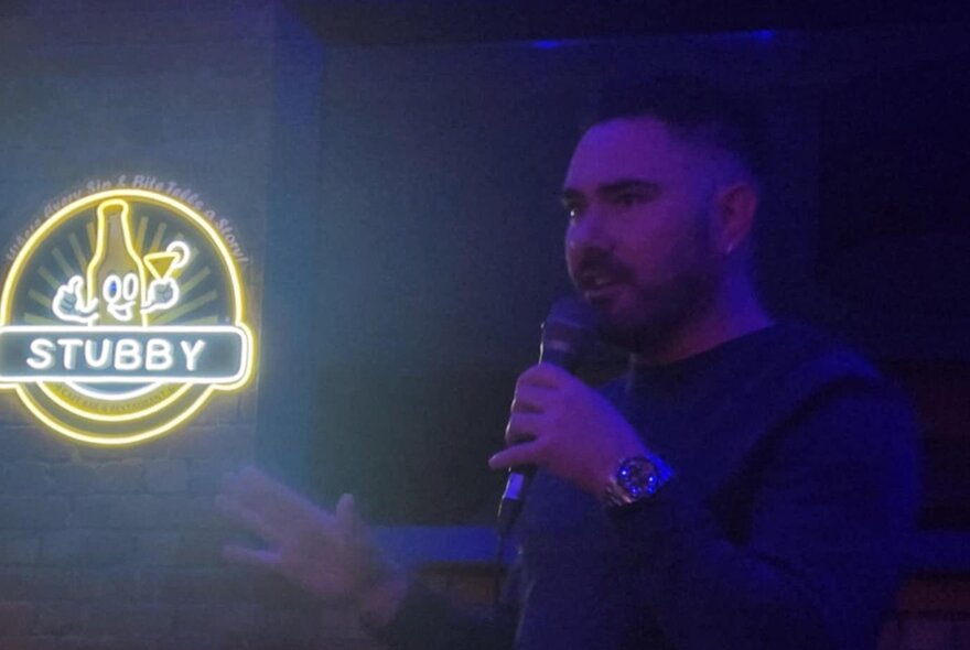 A comedian on a dark stage holding a microphone with a neon sign saying STUBBY and a cartoonish beer bottle smiling. 