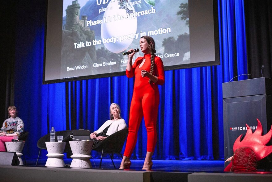 A woman in a red unitard talking into a micriphone while on a stage with a projector screen and blue curtain behind her.