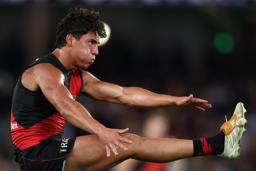 An Essendon AFL football player with arms outstretched during a game and leg raised in the air after having kicked the football.