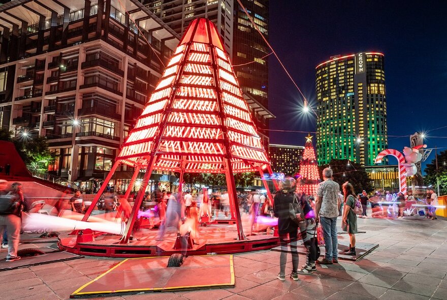 People standing around giant Christmas decorations with koalas climbing lit-up candy canes at Queensbridge Square at night, city buildings in the background.