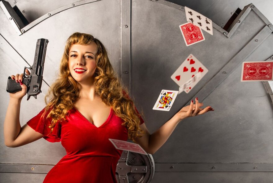 Performer Magical Katrina, wearing a red dress and flicking a pack of oversize playing cards into the air, holding a prop gun. 