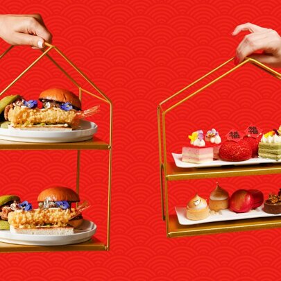 Two hands holding tiered trays of delicate high tea snacks and desserts with a red pattered backdrop behind.