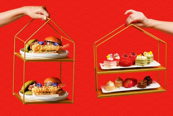 Two hands holding tiered trays of delicate high tea snacks and desserts with a red pattered backdrop behind.