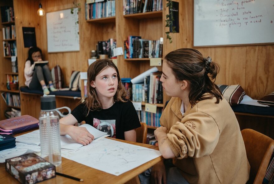 Two children in a studio room, seated at a work table, interacting with one another, an open notepad in front of them, with bookshelves in the background.