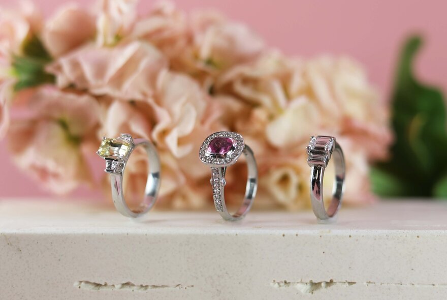 Three diamon and precious stone rings balanced on their sides with blurred pink flowers in the background. 