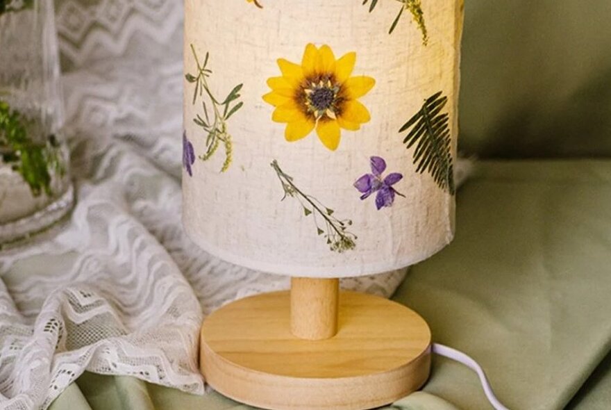 A lamp with a cylindrical parchment or fabric shade, decorated with various pressed flowers, on a small table.
