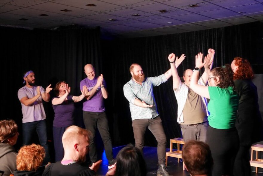 Seven actors on stage performing and clapping, in front of an audience.