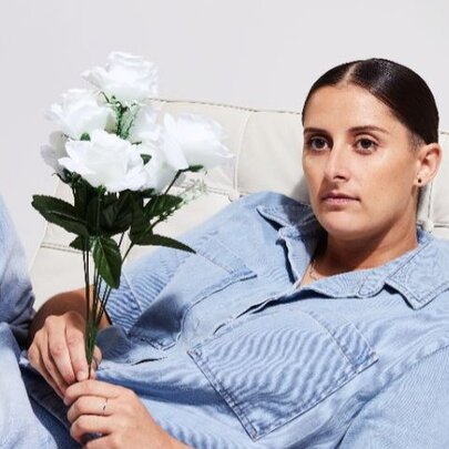 Comedian Annie Boyle wearing a denim shirt and holding white roses in her hand, reclining and with a neutral expression on her face.