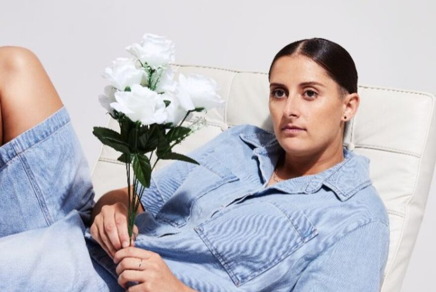 Comedian Annie Boyle wearing a denim shirt and holding white roses in her hand, reclining and with a neutral expression on her face.