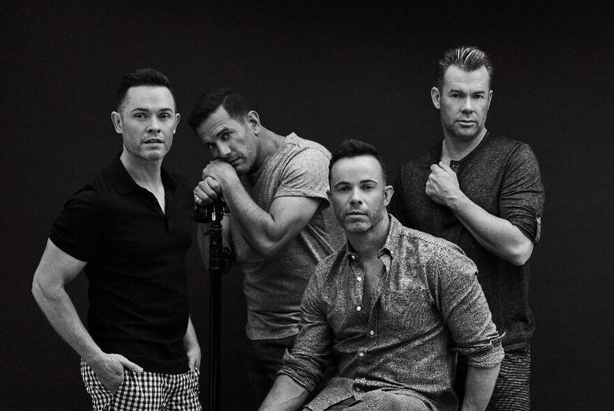 Four-piece musical group of men wearing casual shirts against black background.