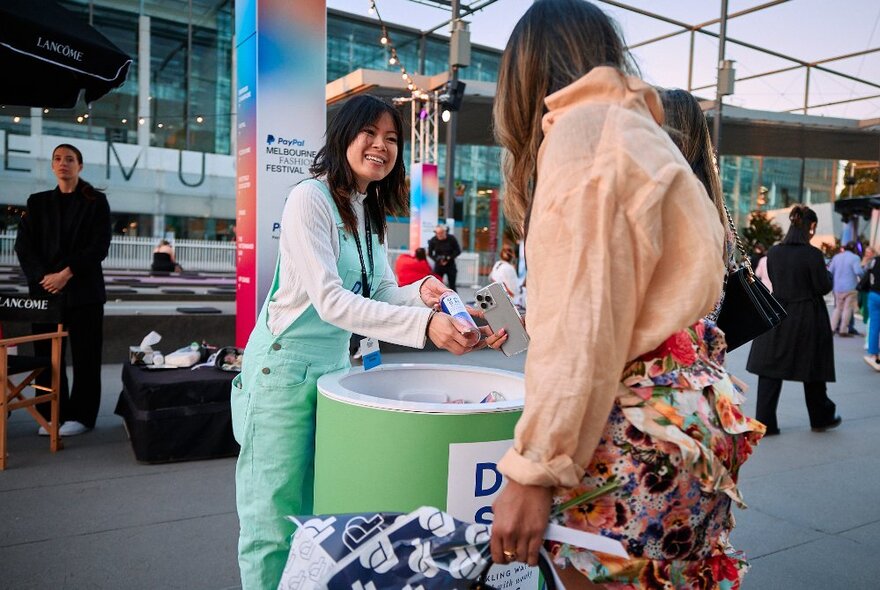 Two people interacting in Museum Plaza, one person handing the other person a bottled drink from a large tub, with people wandering in the background.