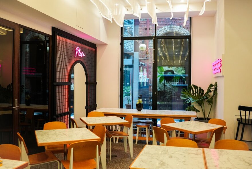 Cafe interior with small wooden tables and chairs, pink neon signage and street-facing window.