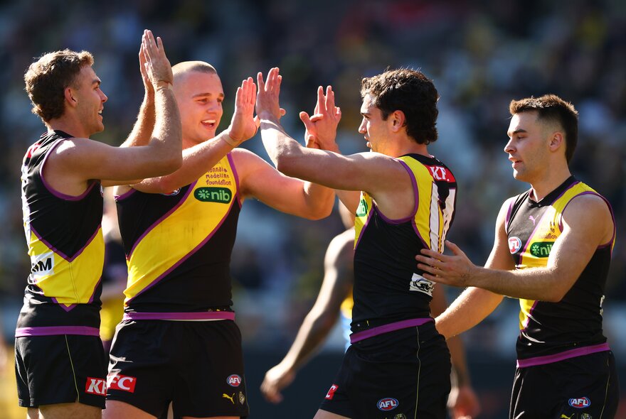 Richmond AFL players in a huddle celebrating on the field after having scored, they face each other and are high-fiving their hands.