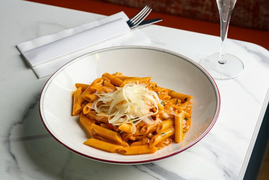 A plate of penne pasta with a tomato sauce and shaved parmesan, on a marble table.