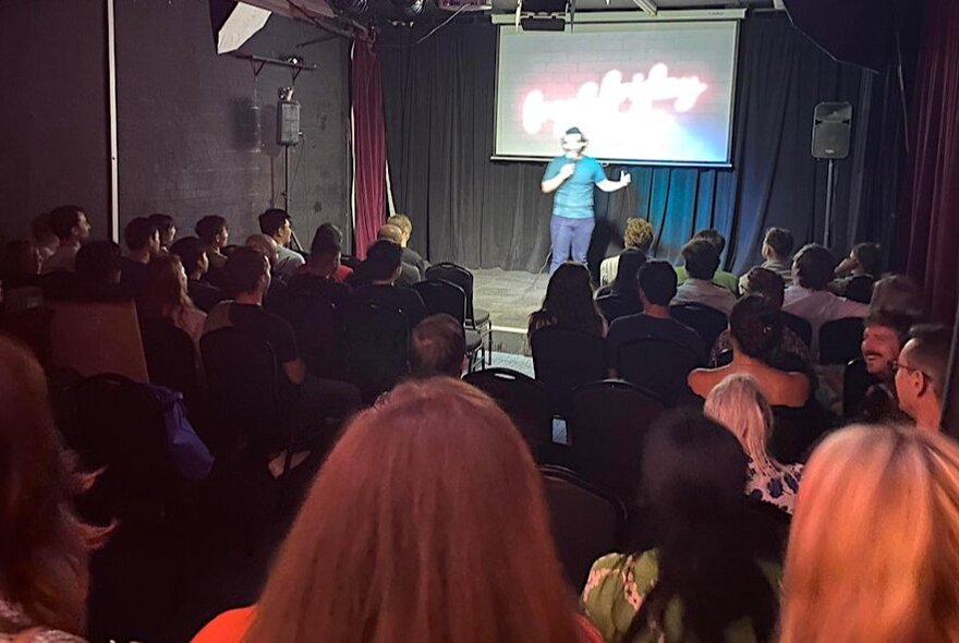 A comedian performing stand-up on a stage, holding a microphone, in front of a seated audience.