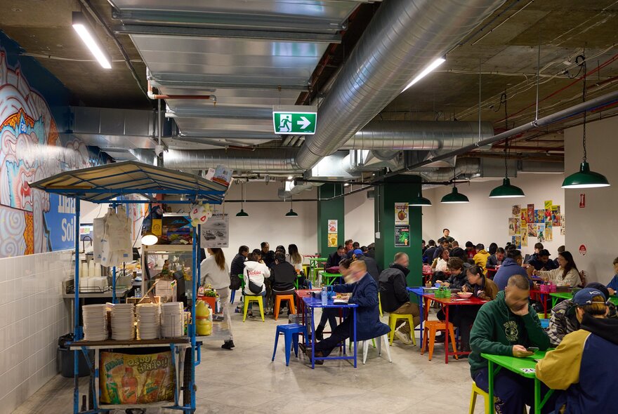 A crowd of people are dining inside a Thai restaurant there are colourful stools and tables