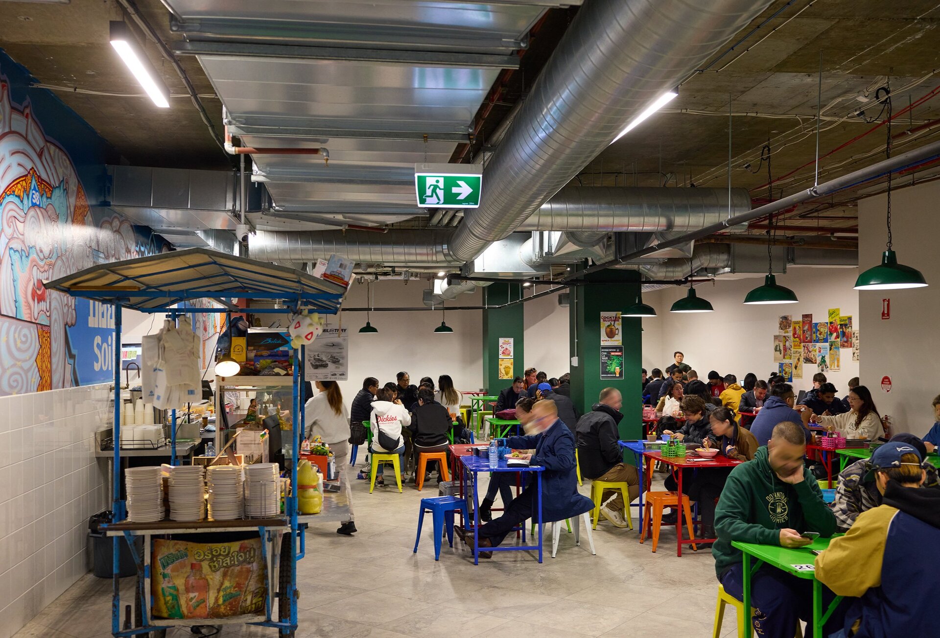 A crowd of people are dining inside a Thai restaurant there are colourful stools and tables