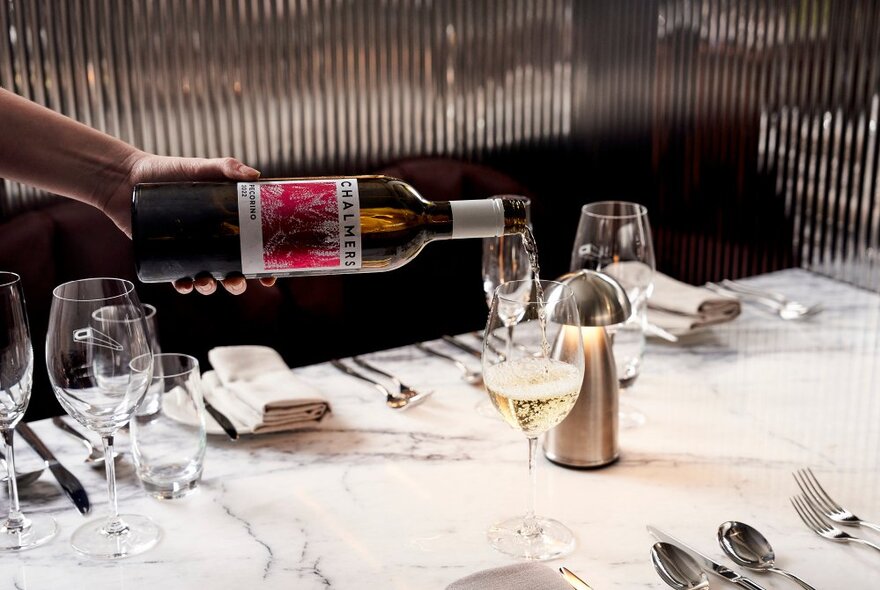 A hand holding a bottle, pouring a glass of wine at a marble table set with cutlery and napkins.