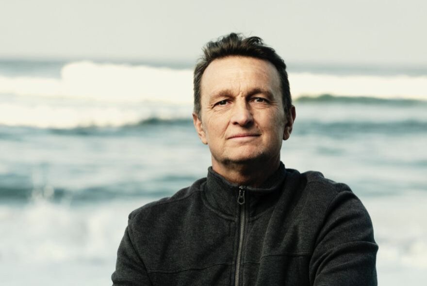Person standing with waves crashing behind them, wearing a dark windcheater.