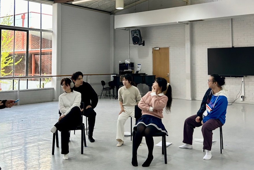 People seated on chairs in a light venue with windows and white painted floor.