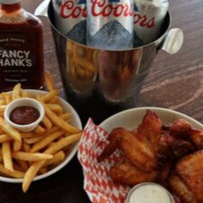 A bowl of spicy chicken wings, chips with tomato sauce and a bottle of Fancy Hanks sauce and Coors beers.