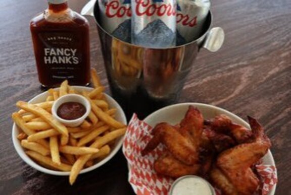 A bowl of spicy chicken wings, chips with tomato sauce and a bottle of Fancy Hanks sauce and Coors beers.