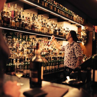 A bartender selecting a bottle from shelves of whiskies and other bottes behind a bar counter.