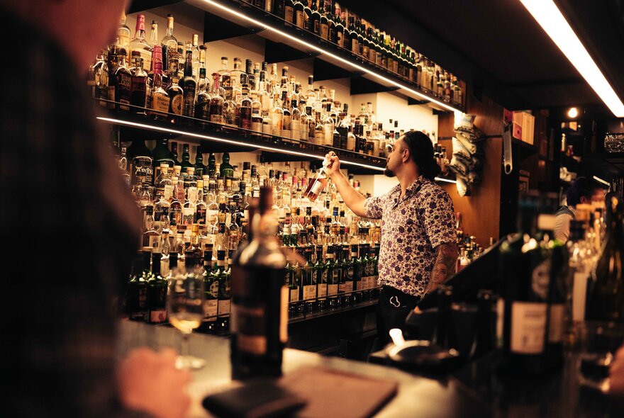 A bartender selecting a bottle from shelves of whiskies and other bottes behind a bar counter.