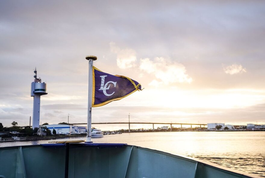 The bow of a boat with a flag flying with the initials LC on it, and a view of the harbour and sky and a bridge in the far distance.