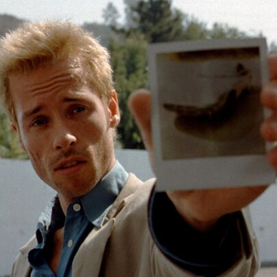 A still from the film Memento showing the actor Guy Pearce with a blond buzz cut and holding up a polaroid. 