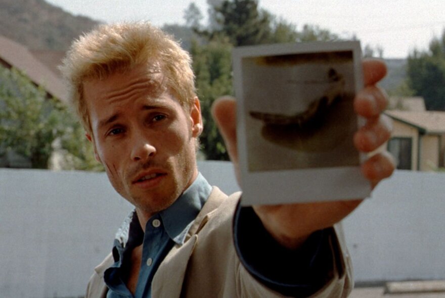 A still from the film Memento showing the actor Guy Pearce with a blond buzz cut and holding up a polaroid. 