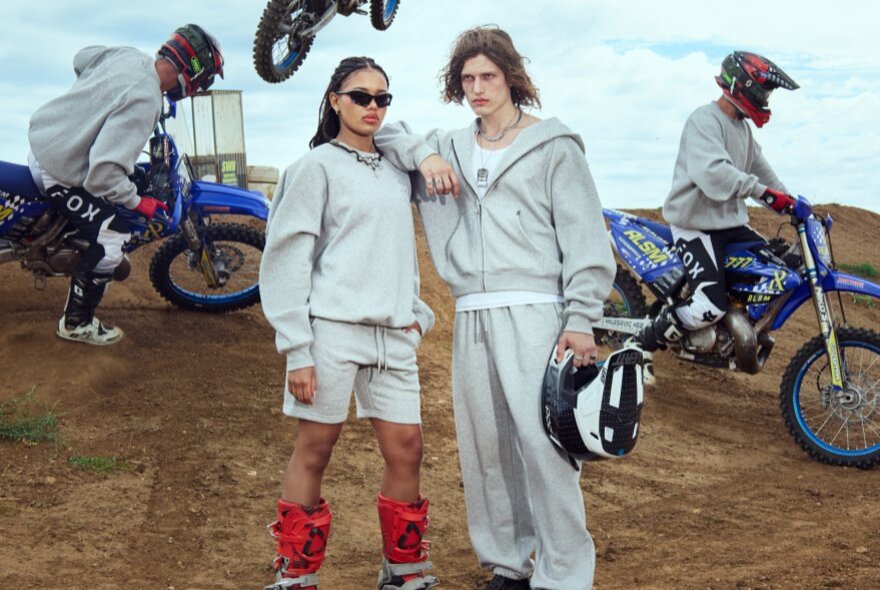 Two models wearing hoodies, trackpants and shorts standing in a field with dirt bike riders in the background.
