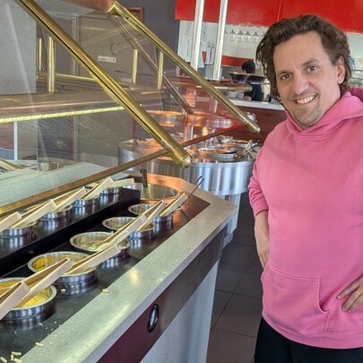 Comedian Ben Russell wearing a bright pink hoodie smiling as he stands next to a buffet-style food station with several metal bowls and serving utensils under a glass cover.