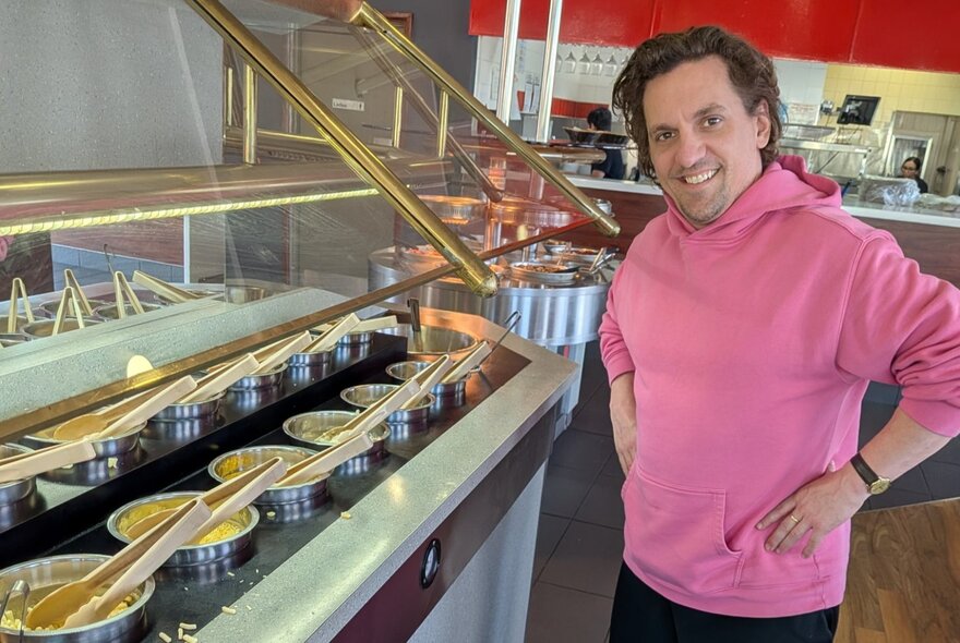 Comedian Ben Russell wearing a bright pink hoodie smiling as he stands next to a buffet-style food station with several metal bowls and serving utensils under a glass cover.
