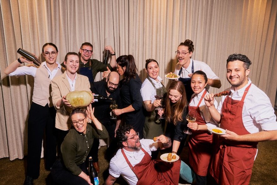 A group of hospitality people in aprons and uniforms casually posing for a group shot and laughing. 