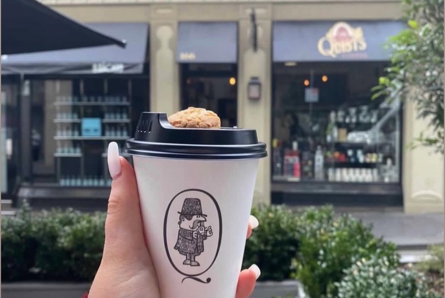 A hand holding a takeaway cup of coffee in the foreground, with Quists Coffee shop in Little Collins Street slightly out of focus in the background.