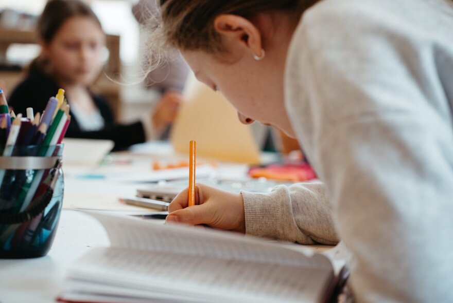 A person leaning over and writing in a notebook at a communal desk. 