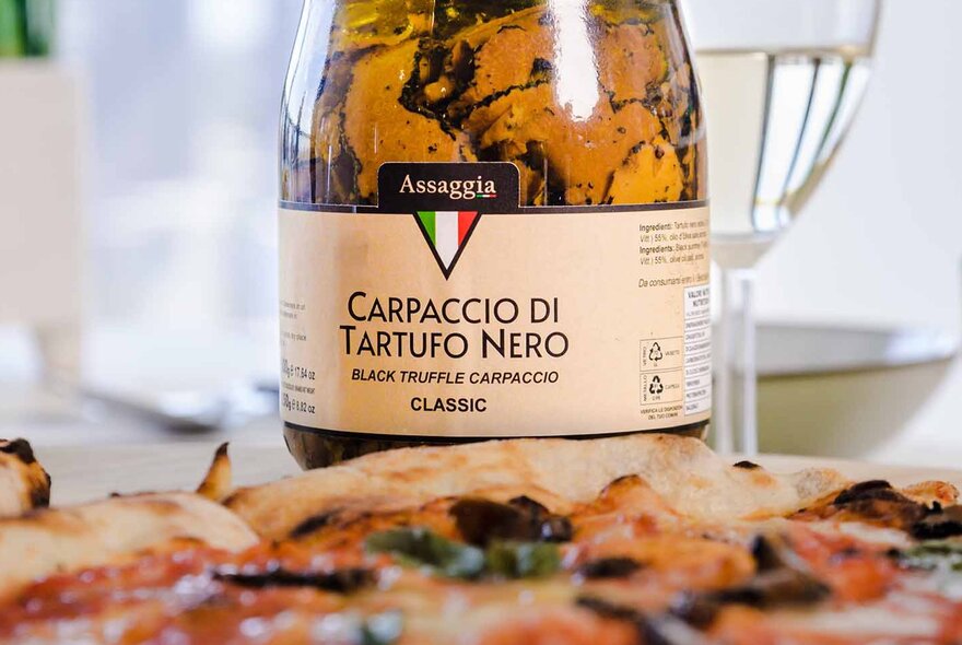 A jar of carpaccio di tartufo nero (thinly sliced black truffle); a woodfired Italian pizza in the foreground.